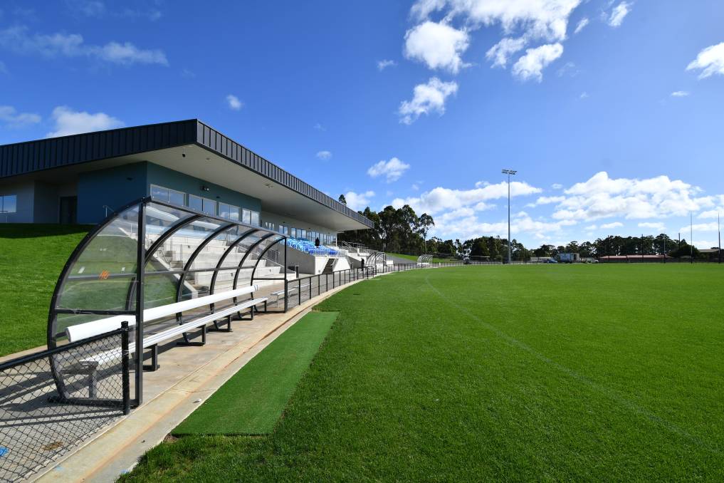 View along the boundary line at Dial Park in Penguin, showing the main grandstand, player bench shelter, green playing surface, and lighting towers