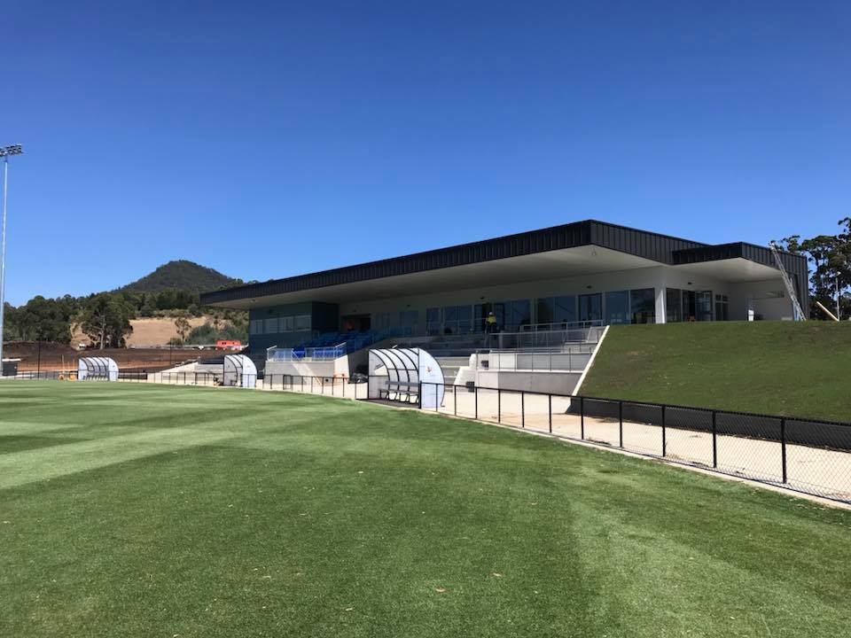 View of the main grandstand and player shelters at Dial Park in Penguin, with tiered seating, glass-fronted clubhouse facilities, and the playing field in the foreground