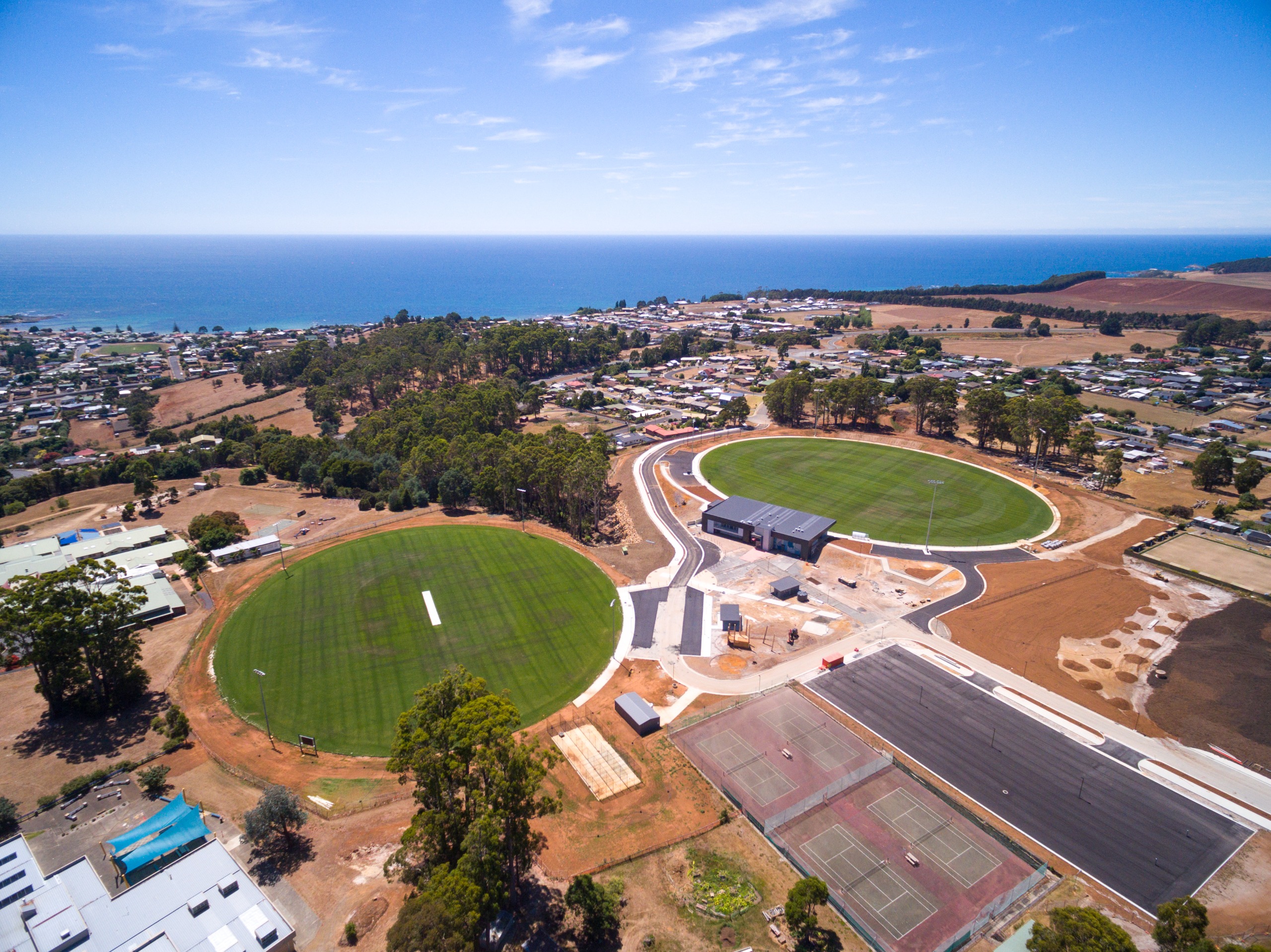 Aerial view of the Dial Sports Complex in Penguin, Tasmania, showing two large green ovals, a central clubhouse, tennis courts, and surrounding community facilities, with the town and coastline in the background.